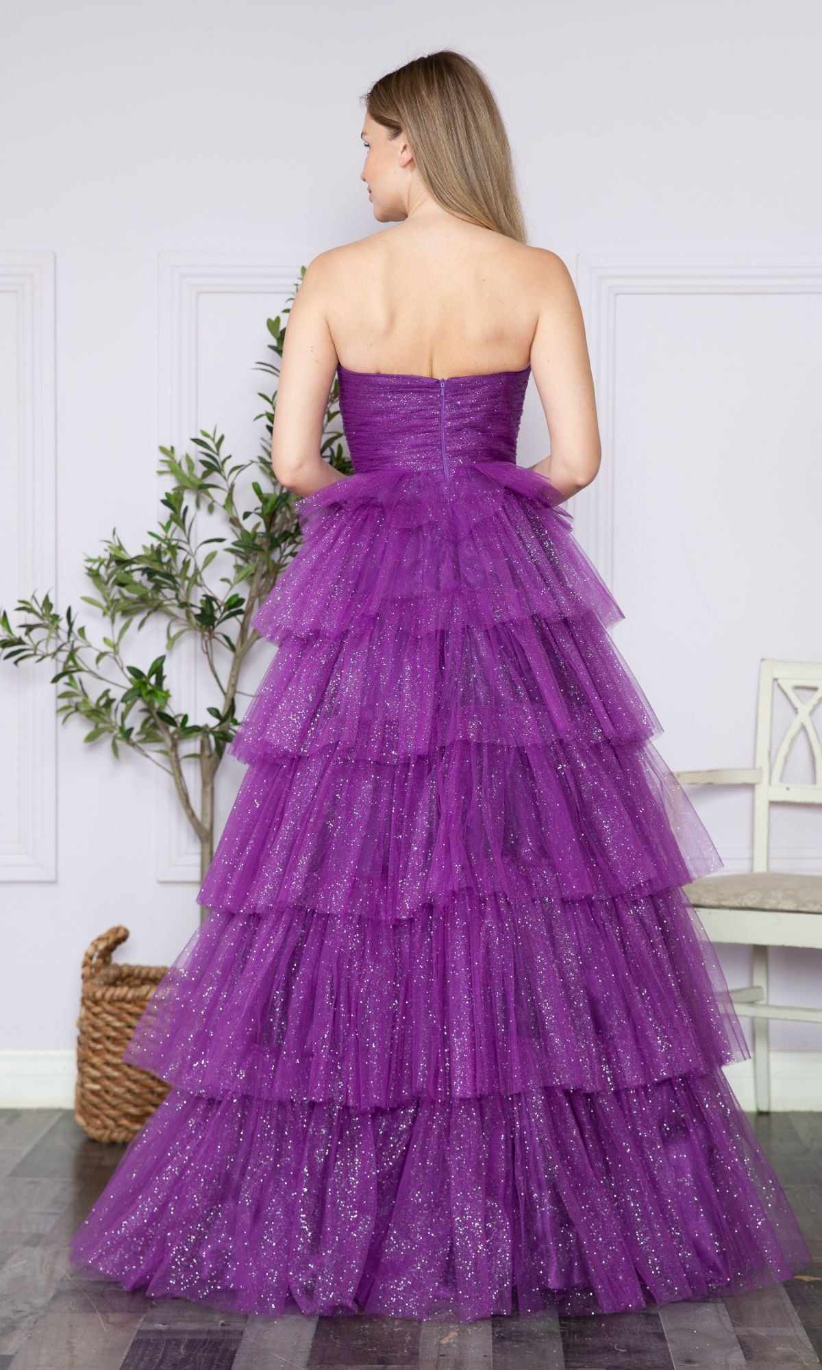 A woman stands indoors facing away, wearing the Strapless Long Tiered Glitter Prom Ball Gown 9386—floor-length purple tulle with glitter and tiered layers. A plant and a chair are in the background.